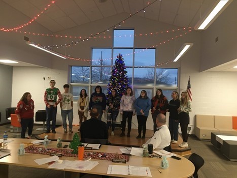 High School choir singing Christmas carols in front of large window and Christmas tree to school board